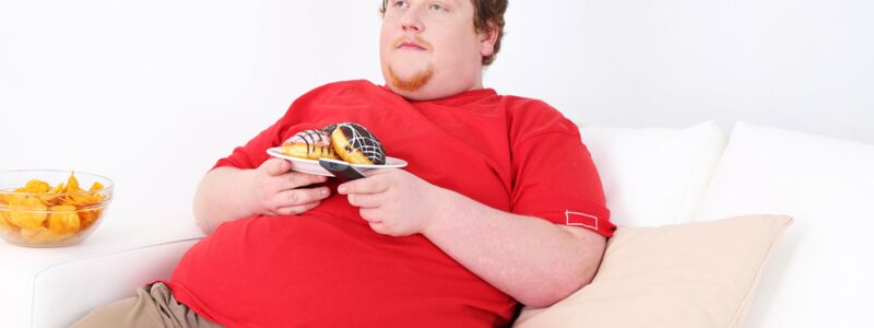 Overweight man sits on the couch and eats chips and doughnuts